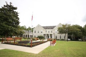 Front view of The Farley Center, a beige two-story building with white trim and columns, surrounded by a well-maintained lawn, red benches, landscaped flowerbeds, and a central walkway leading to the entrance. An American flag is prominently displayed on a flagpole in the courtyard.