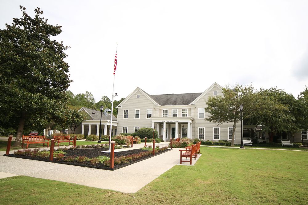 Front view of The Farley Center, a beige two-story building with white trim and columns, surrounded by a well-maintained lawn, red benches, landscaped flowerbeds, and a central walkway leading to the entrance. An American flag is prominently displayed on a flagpole in the courtyard.