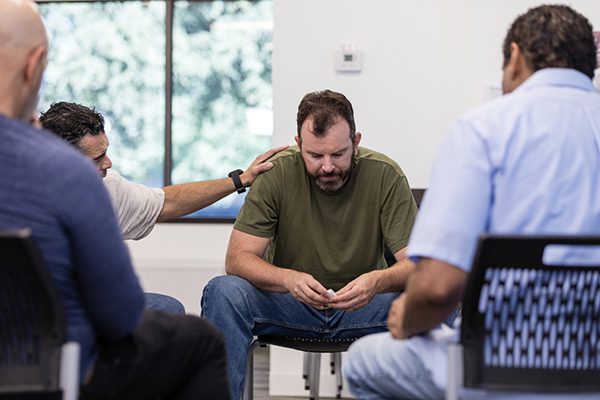 As the other men sit quietly, the mature adult male therapist encourages the emotionally distressed mid adult male veteran.
