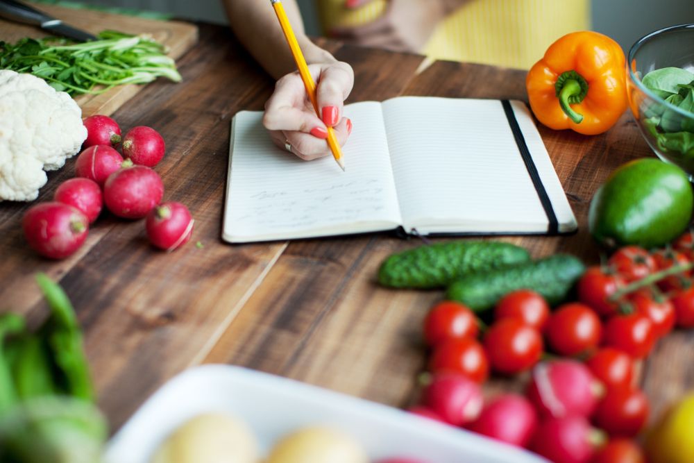 Person writing in a notebook with a pencil on a wooden table surrounded by fresh vegetables, including radishes, peppers, tomatoes, and cauliflower.