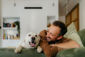 Smiling man relaxing on a couch next to his happy dog, both looking content in a cozy, modern living room.