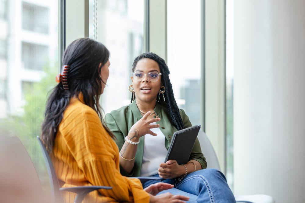 A counselor holding a tablet talks with a woman in an orange sweater during a support session in a bright, modern office.