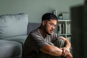 A man sitting on the floor near a couch, looking thoughtful and lonely, symbolizing the struggles of isolation during recovery.