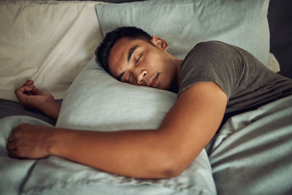 A young man sleeps peacefully on his bed, resting his head on a pillow while wrapped in soft gray bedding.