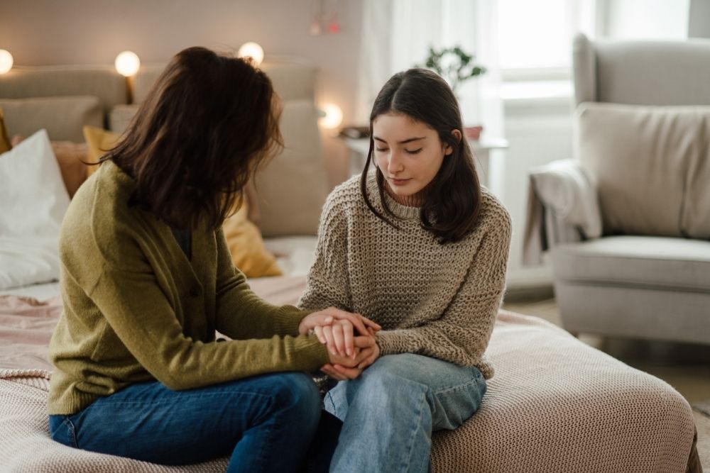 An adult and a young girl sit on a bed holding hands, engaged in a serious and supportive conversation in a cozy, softly lit room.