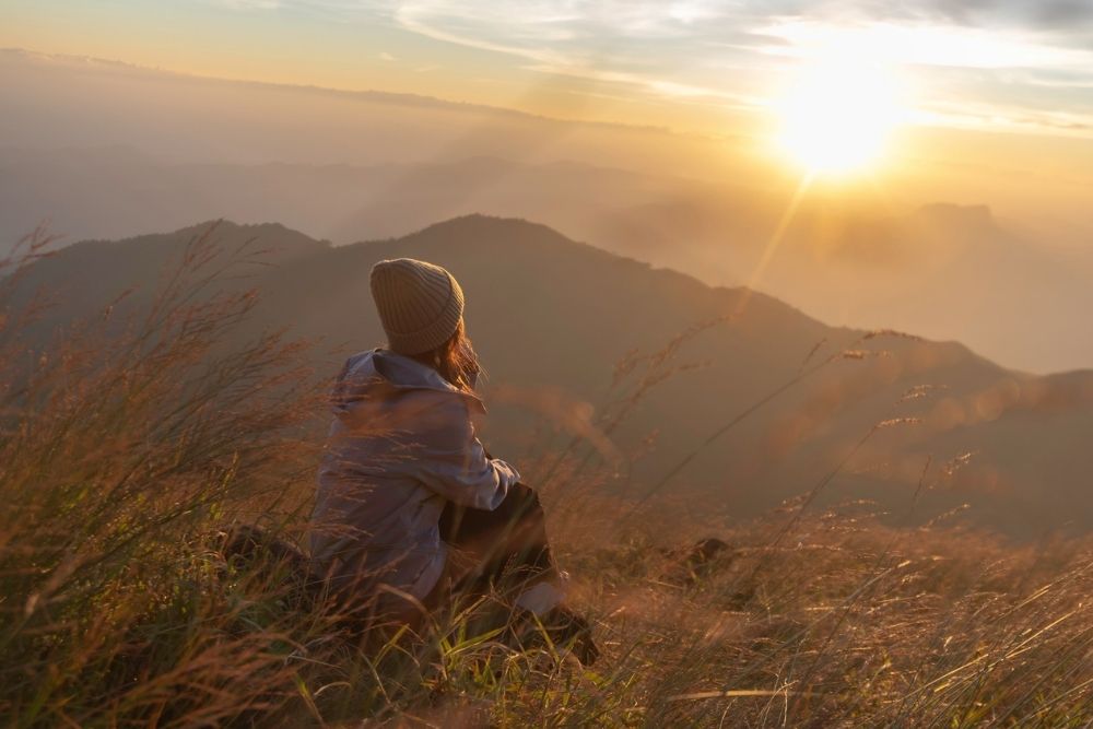Person sitting on a grassy hillside at sunrise, looking out over mountains and valleys, reflecting peacefully in nature.