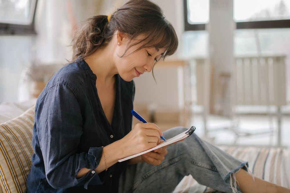 Woman sitting comfortably and writing in a notebook, appearing thoughtful and focused, suggesting reflection or personal growth.
