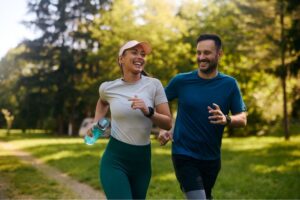 A smiling man and woman jog together through a sunny park, enjoying an outdoor workout surrounded by trees and greenery.