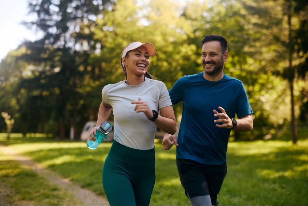 A smiling man and woman jog together through a sunny park, enjoying an outdoor workout surrounded by trees and greenery.
