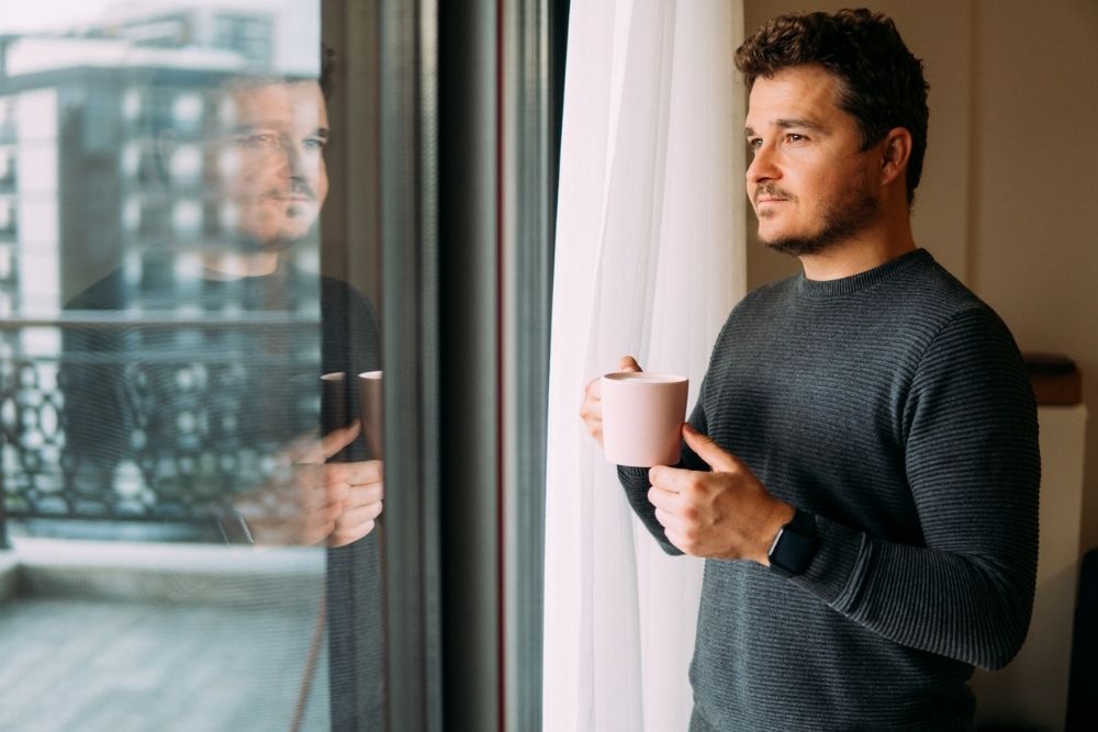 A man stands by a window holding a mug, looking outside with a thoughtful expression, his reflection visible in the glass.
