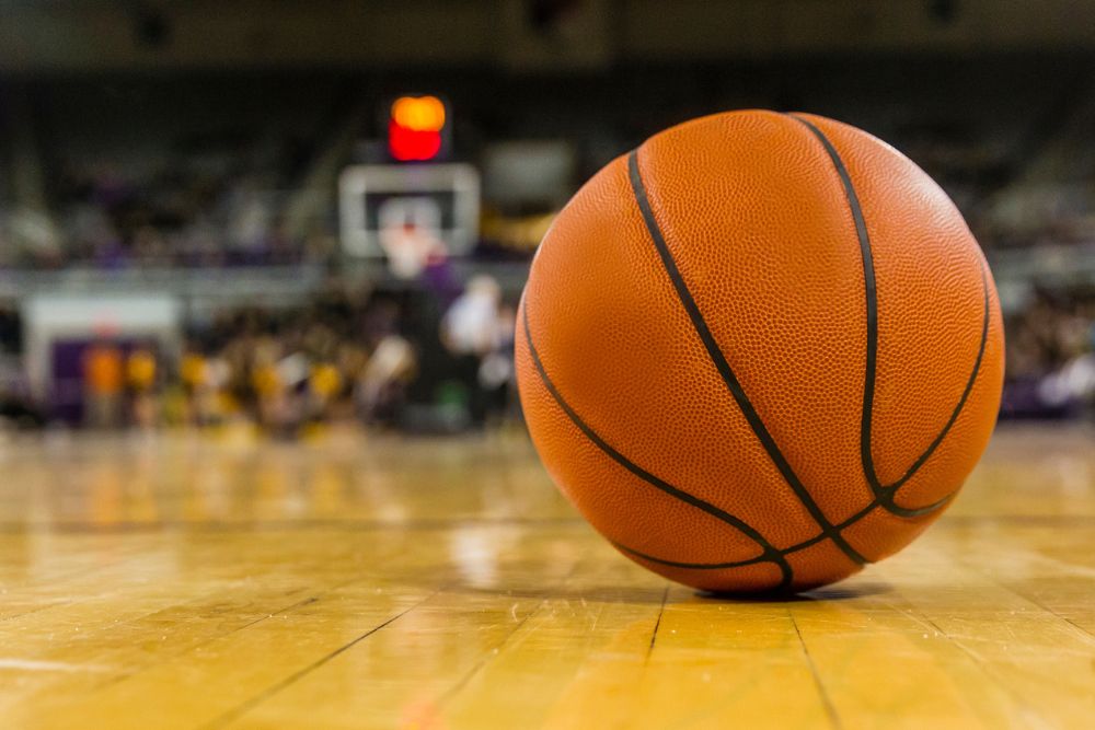 A basketball resting on a polished gym floor with a blurred basketball court and players in the background.