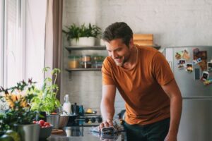 Man smiles while wiping down a kitchen counter, cleaning up in a bright home kitchen with plants nearby.