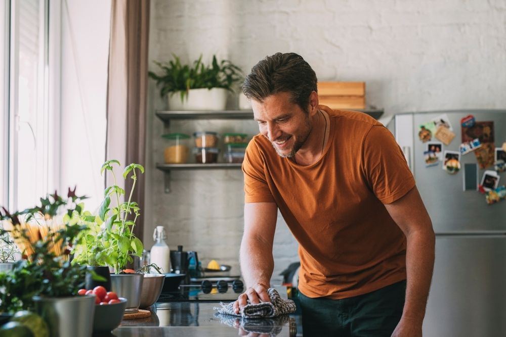 Man smiles while wiping down a kitchen counter, cleaning up in a bright home kitchen with plants nearby.